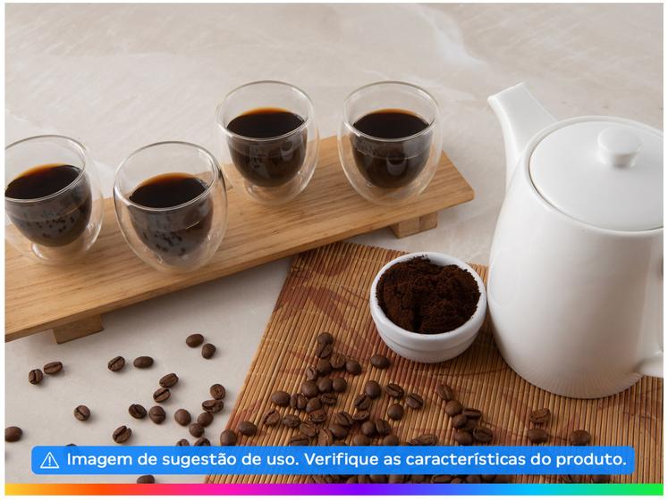 Um conjunto de quatro copos de café de vidro com parede dupla sobre uma bandeja de madeira, ao lado de um bule branco, uma tigela de café moído e grãos de café espalhados sobre uma esteira texturizada.