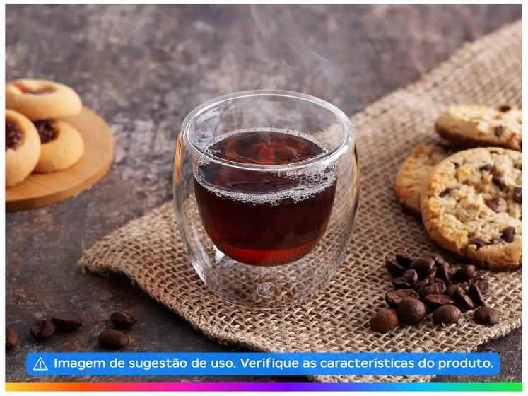 Um único copo de vidro com parede dupla contendo uma bebida escura quente e fumegante, colocado sobre um tecido texturizado com biscoitos e grãos de café ao redor.
