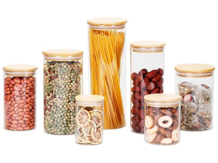 Conjunto de 7 potes de vidro Brinox Ekko, alguns cheios de alimentos como amendoim, ervilhas, macarrão espaguete, jujubas, tâmaras e chá, com tampas de bambu, em fundo branco.